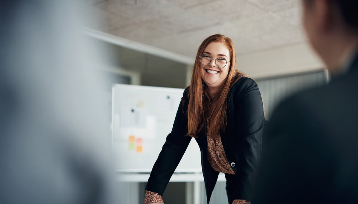Woman smiling while standing at a table in front of her coworkers, with a 50 out of 60 scale and a green ‘Model the Way’ leadership behavior banner are in the bottom right corner.